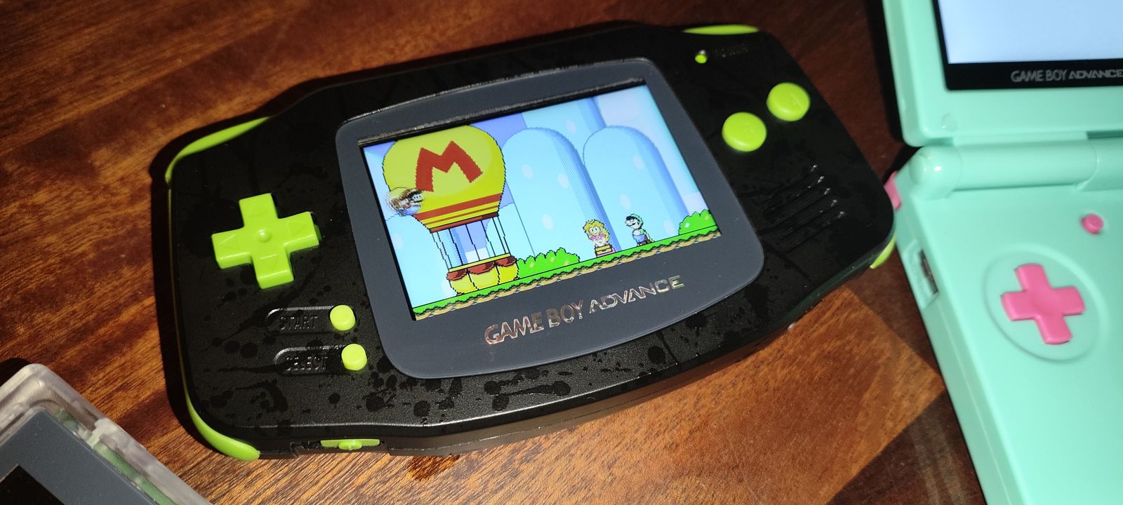 A black splatter Game Boy Advance with lime green buttons and a bright IPS screen running a Mario game