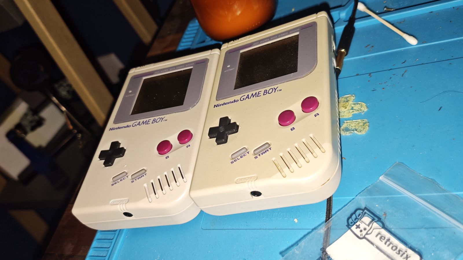 Two original grey Game Boys side by side on a blue silicone work mat, next to a bag of parts
