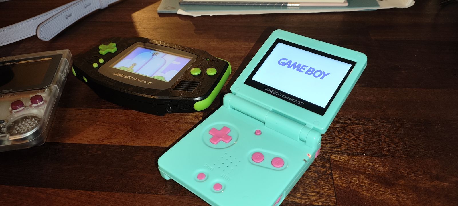 A mint Game Boy Advance SP with pink buttons showing the Game Boy boot screen, next to the black and lime Game Boy Advance